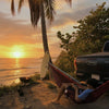 Person relaxing in a hitch hammock by a palm tree on a beach at sunset.