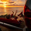 Two people in a hitch hammock by the ocean at sunset, with a car in the foreground.